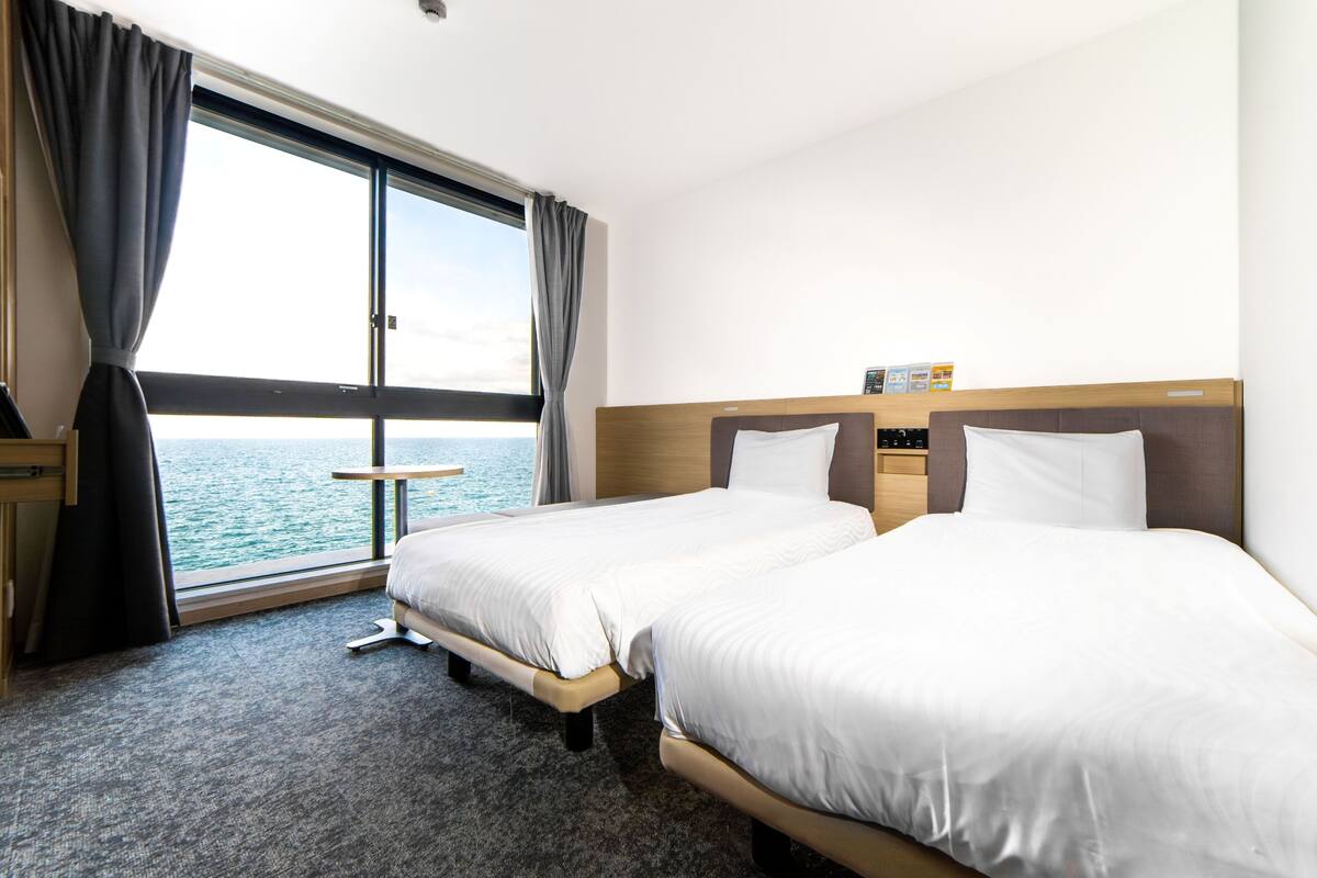 A hotel room with two beds, a small table, and a view of the sea.