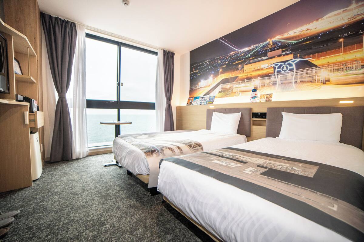 A hotel room with two beds, a large window with curtains, a desk with a chair, and a wall-mounted TV.