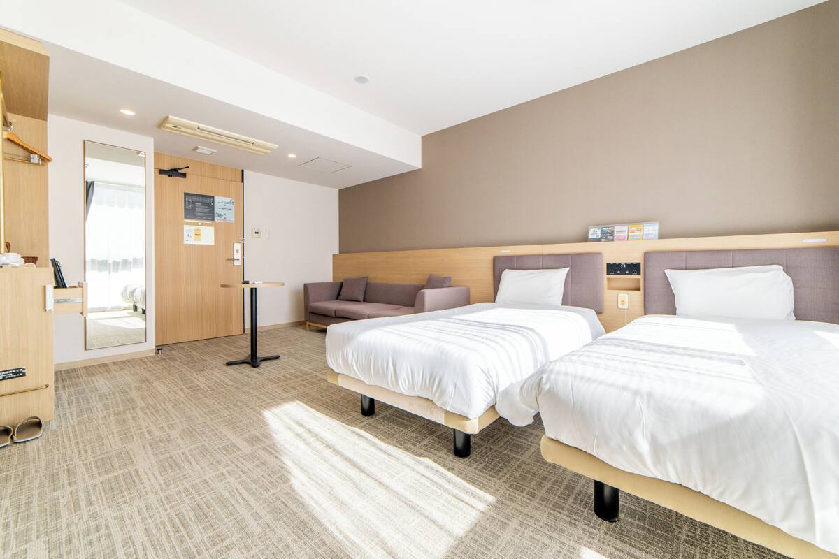 A hotel room with two beds, a sofa, a mirror, and a shoe rack.