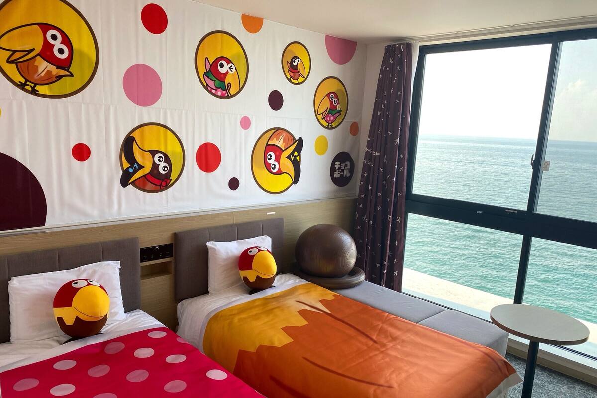 A hotel room with a bed, pillows, a round table, and a large window with a view of the sea.