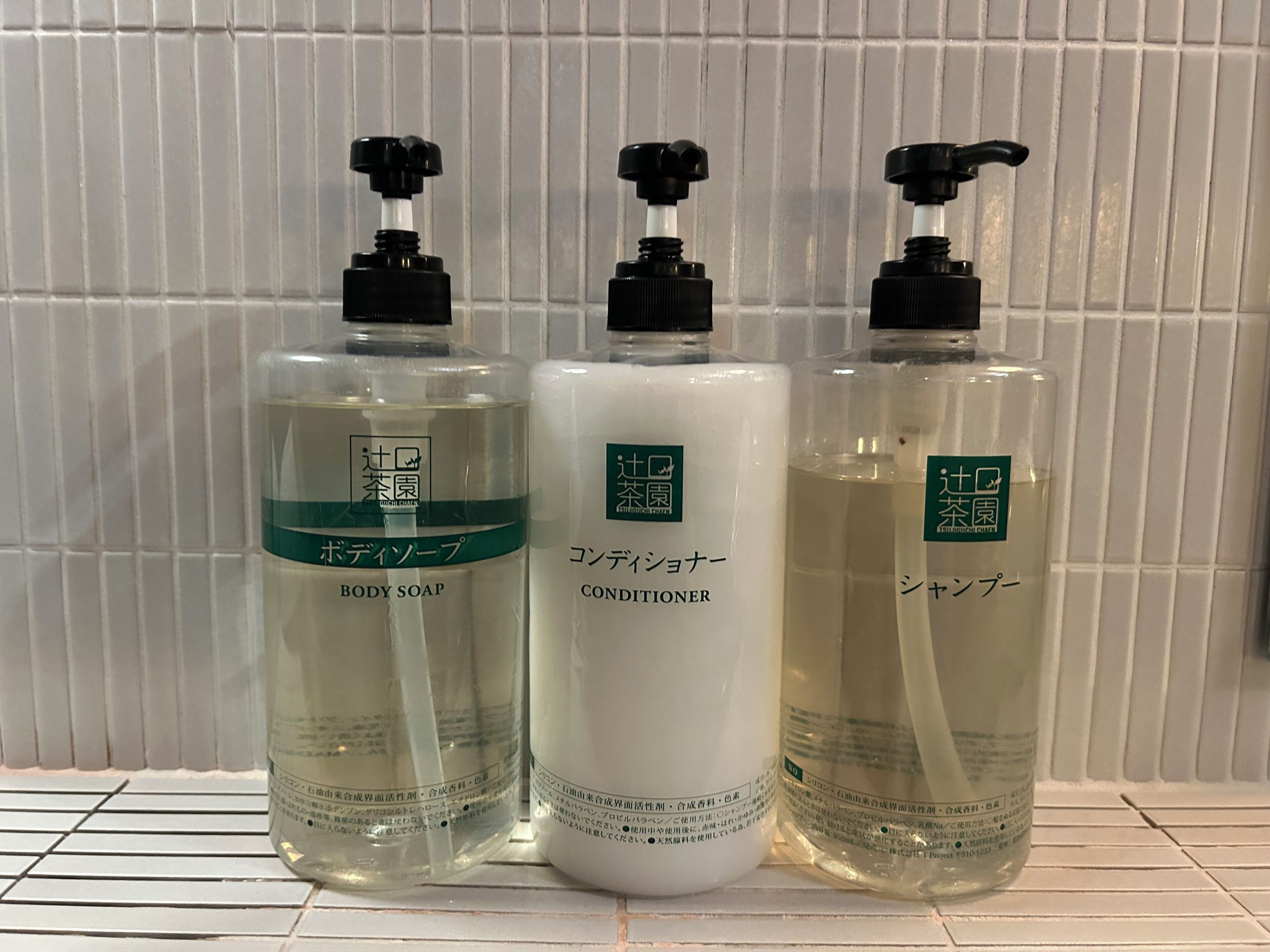 Three bottles of liquid products with pump dispensers, labeled in a foreign language, on a tiled shelf.