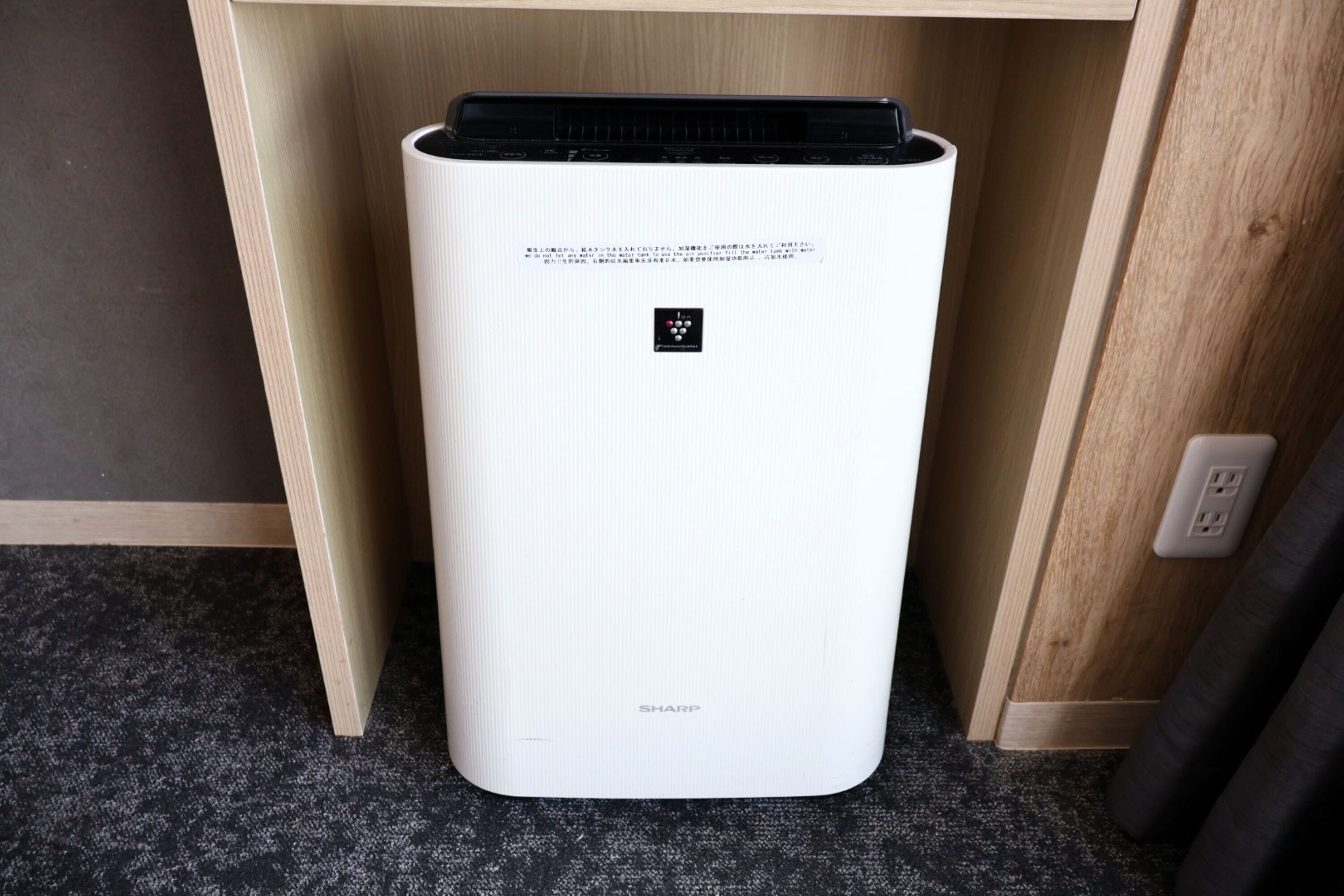 A white air purifier with a black top, placed under a wooden shelf in a room with a carpeted floor.
