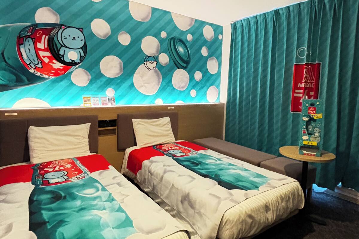 A hotel room with two beds, a mural with cartoon characters, a small table with a red sign, and teal curtains.