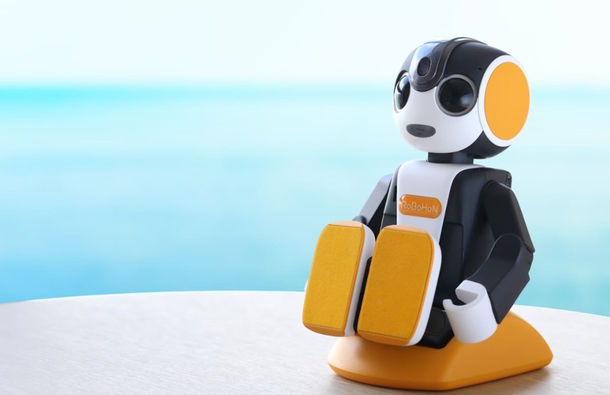 Un robot con la etiqueta 'RoBoHoN' en el pecho, sentado sobre una base amarilla.