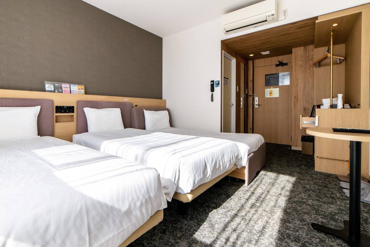 A hotel room with two beds, a desk, a wardrobe, and an air conditioning unit.