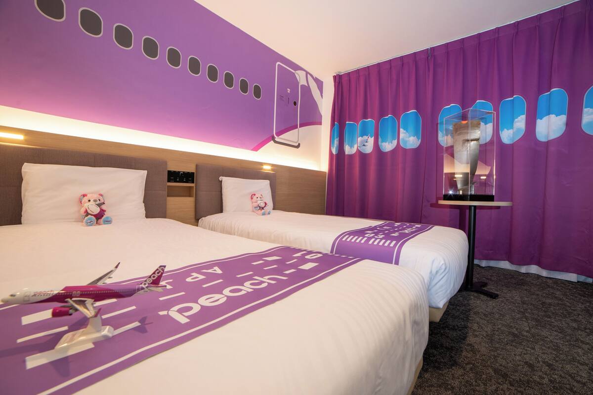 A hotel room with two beds, a large window with purple curtains, a wall-mounted TV, and a small table.