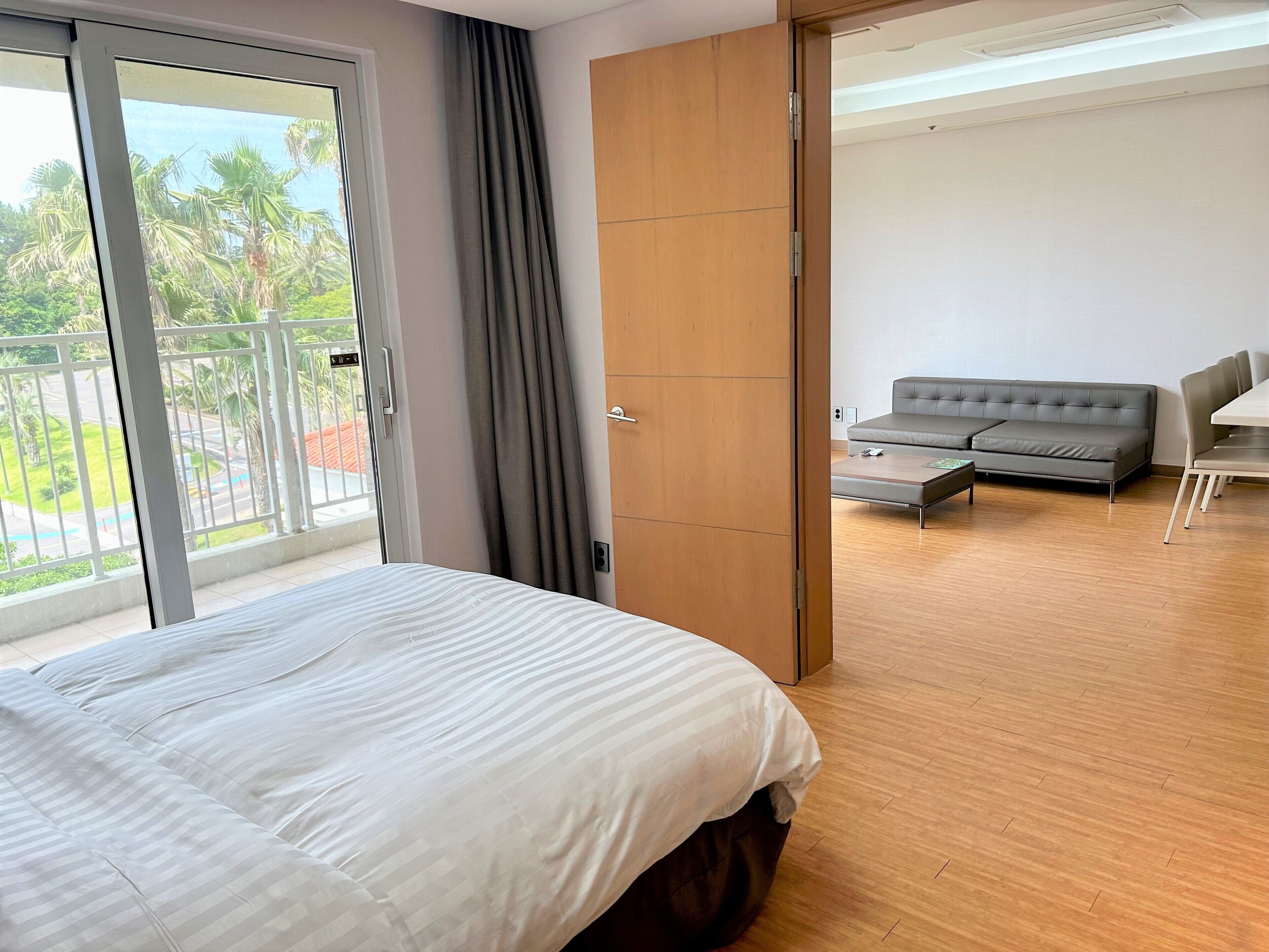 Une chambre d’hôtel comprenant un lit, un canapé, une table à manger et une porte coulissante donnant sur un balcon avec vue sur des palmiers et une piscine.