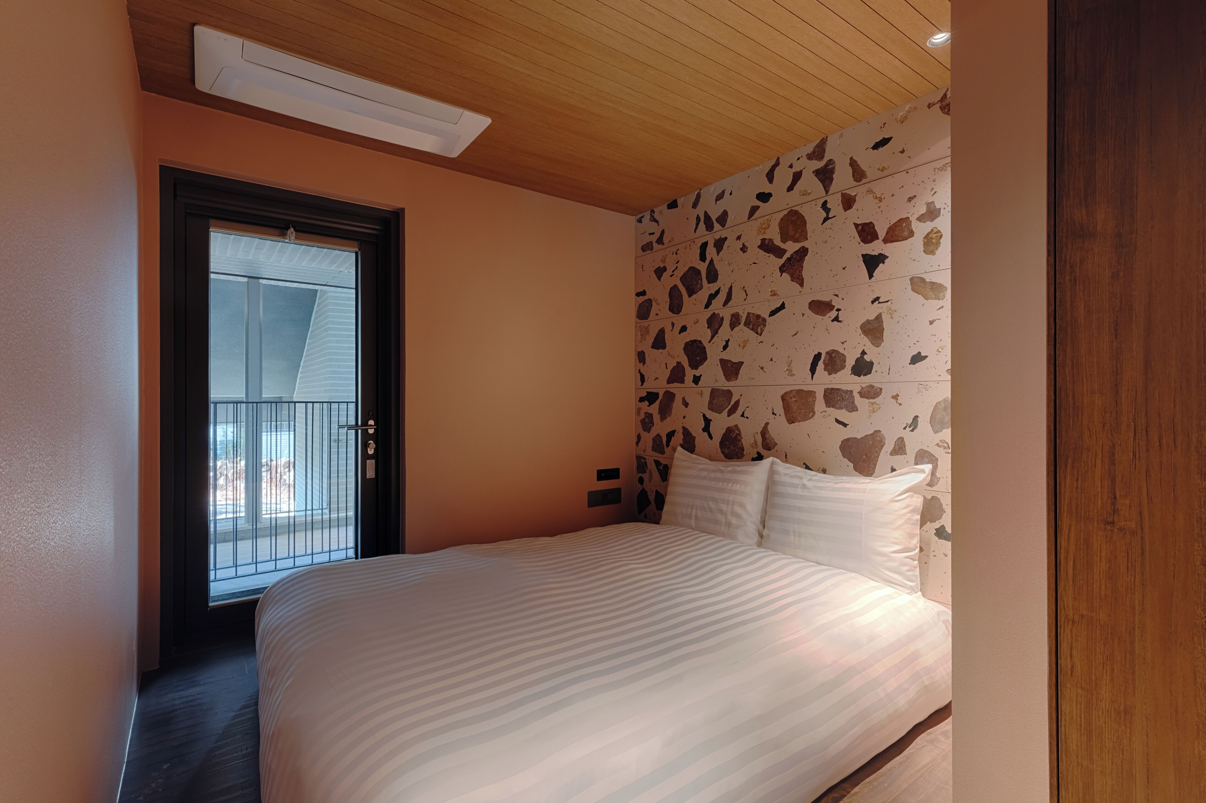 Een slaapkamer met een bed, een raam met zonwering, een muur met steenmotief en een houten plafond.