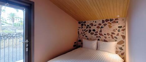 A bedroom with a large bed, a stone-patterned headboard, and a sliding glass door leading to an outdoor area with a stone wall and palm trees.