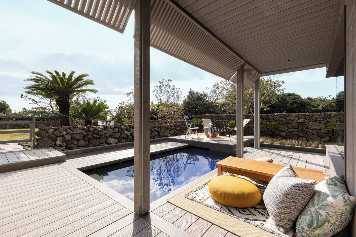 Ein Poolbereich mit Holzterrasse, Steinmauer und Sitzgelegenheit mit Bank und Kissen.