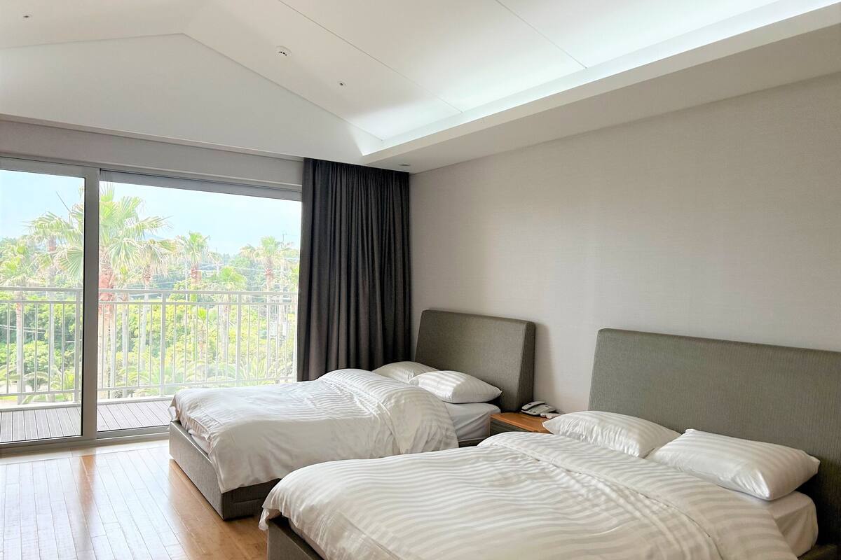 Ein modernes Hotelzimmer mit zwei Betten, einem großen Balkon mit Blick auf Palmen und einer Decke mit Einbauleuchten.