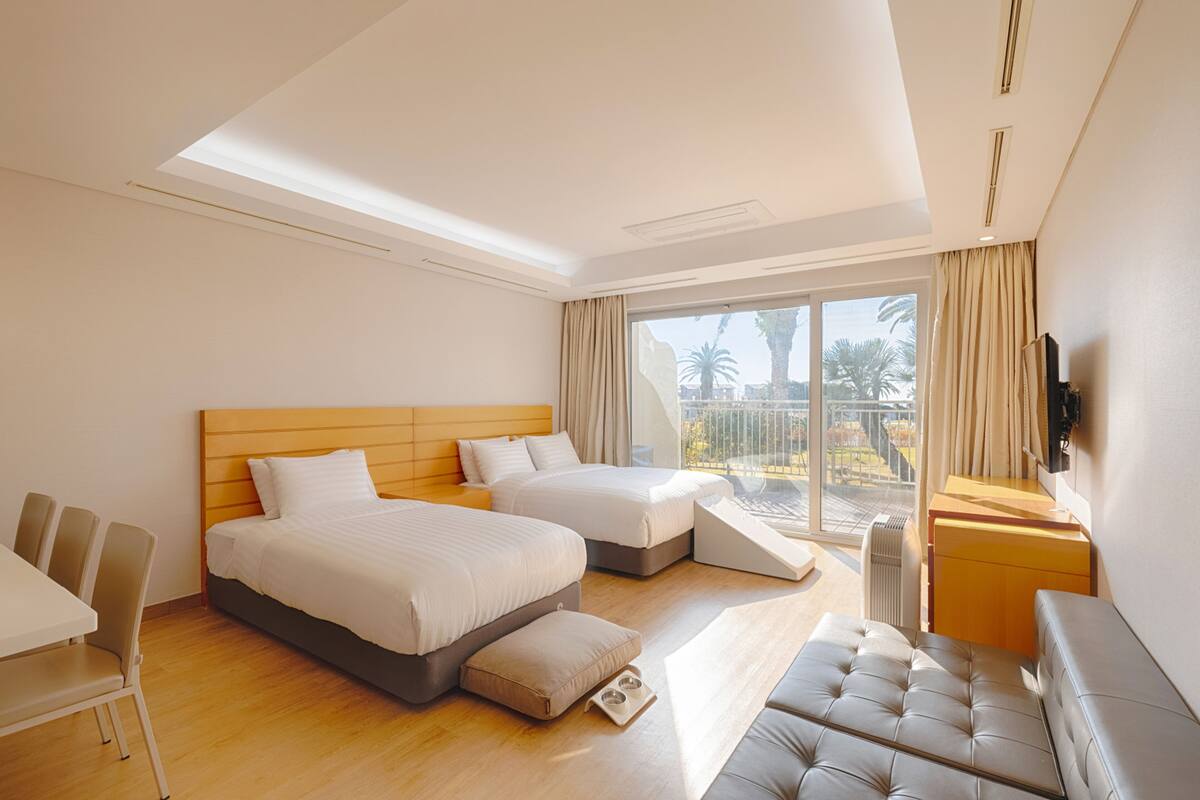Ein modernes Hotelzimmer mit zwei Betten, einer Couch, einem Esstisch und einem Balkon mit Blick auf Palmen.