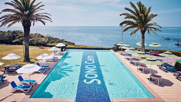 A pool with 'SONO Cam' written on the water, surrounded by lounge chairs and umbrellas, with palm trees and a view of the sea. - Sono Calm Jeju (Seogwipo)