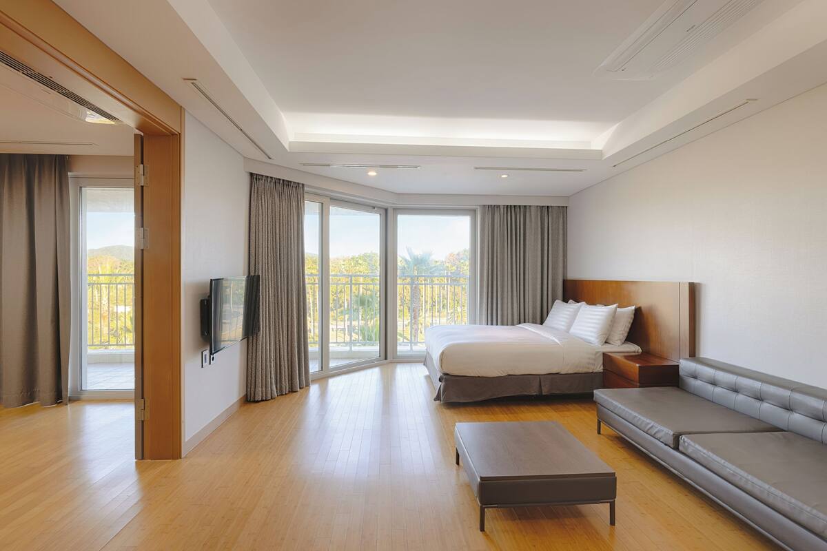 Ein modernes Hotelzimmer mit einem großen Bett, einer Sitzecke, einem Fernseher und einem Balkon mit Blick auf Bäume.