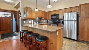 A kitchen with wooden cabinets, a stainless steel refrigerator, a microwave, and a bar area with stools. - Settler's Crossing #37 | True Ski-In/Ski-Out (Sun Peaks)