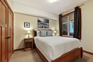 A bedroom with a wooden bed, a nightstand with a lamp, a framed picture on the wall, and a window with curtains. - Incredible ski in/ski out location SX# 37 (EV charger) (Sun Peaks)