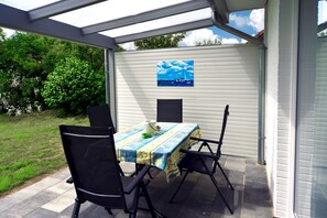Un patio al aire libre con una mesa y sillas, un cuadro de un velero en la pared, y vistas a un jardín y árboles.