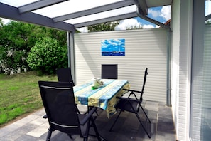 An outdoor patio with a table and chairs, a painting of a sailboat on the wall, and a view of a garden and trees.