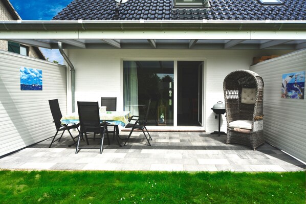 En terrasse med et bord og stole, en fletstol og en skydedør, der fører til et overdækket område.