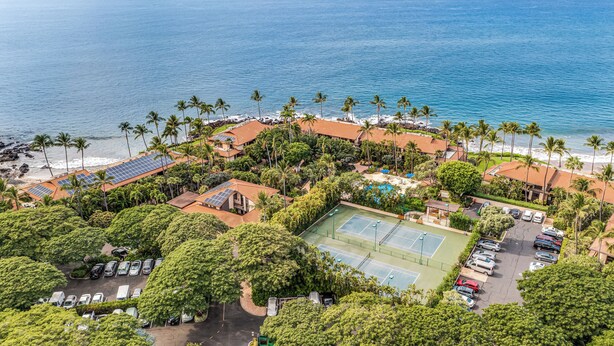 Vue aérienne d’un complexe hôtelier avec une plage, des palmiers, des panneaux solaires sur les bâtiments, un court de tennis et un parking.