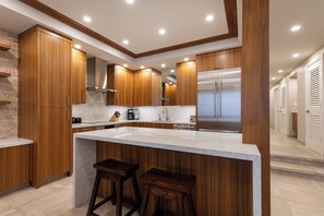 A modern kitchen with wooden cabinets, a large island with stools, and a stainless steel refrigerator. - Exquisite Oceanfront Retreat - Makena Surf F-209 with Stunning Coastal Views! (Makena, Maui)