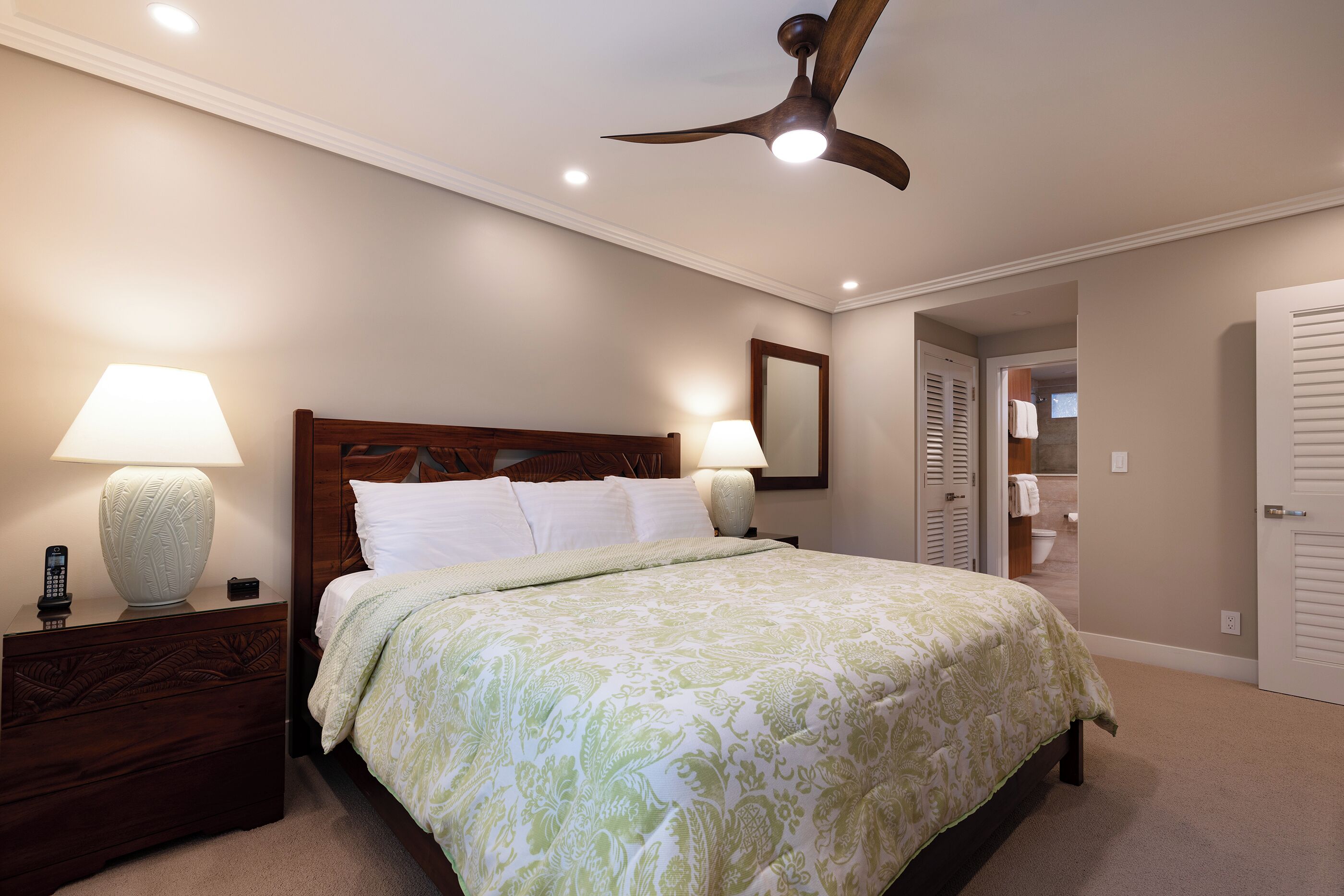 A bedroom with a bed, nightstands, lamps, a ceiling fan, and a door leading to a bathroom.