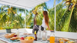 Un coin petit-déjeuner installé sur une terrasse, avec une table recouverte d’une nappe à rayures, où l’on trouve du pain, des fruits et des boissons.