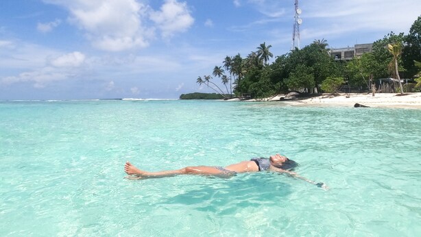 Une personne flotte dans une eau turquoise et limpide, avec une plage et des palmiers en arrière-plan.
