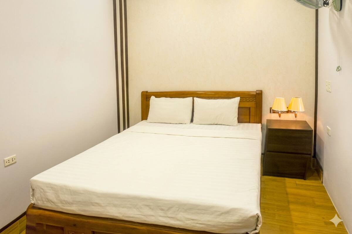 A single bed with a wooden headboard, white bedding, a bedside table with two lamps, and a power outlet on the wall.