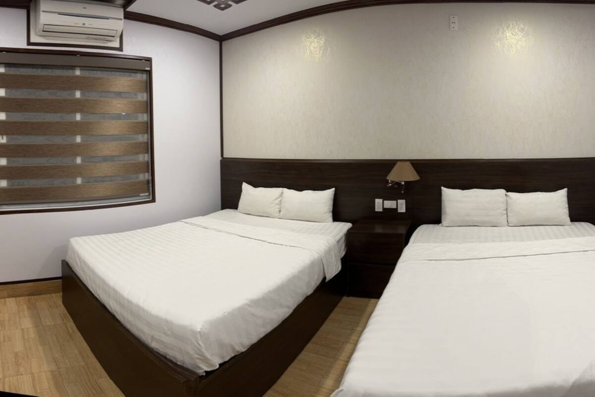 A hotel room with two beds, a window with blinds, a wall-mounted air conditioner, and a nightstand with a lamp.