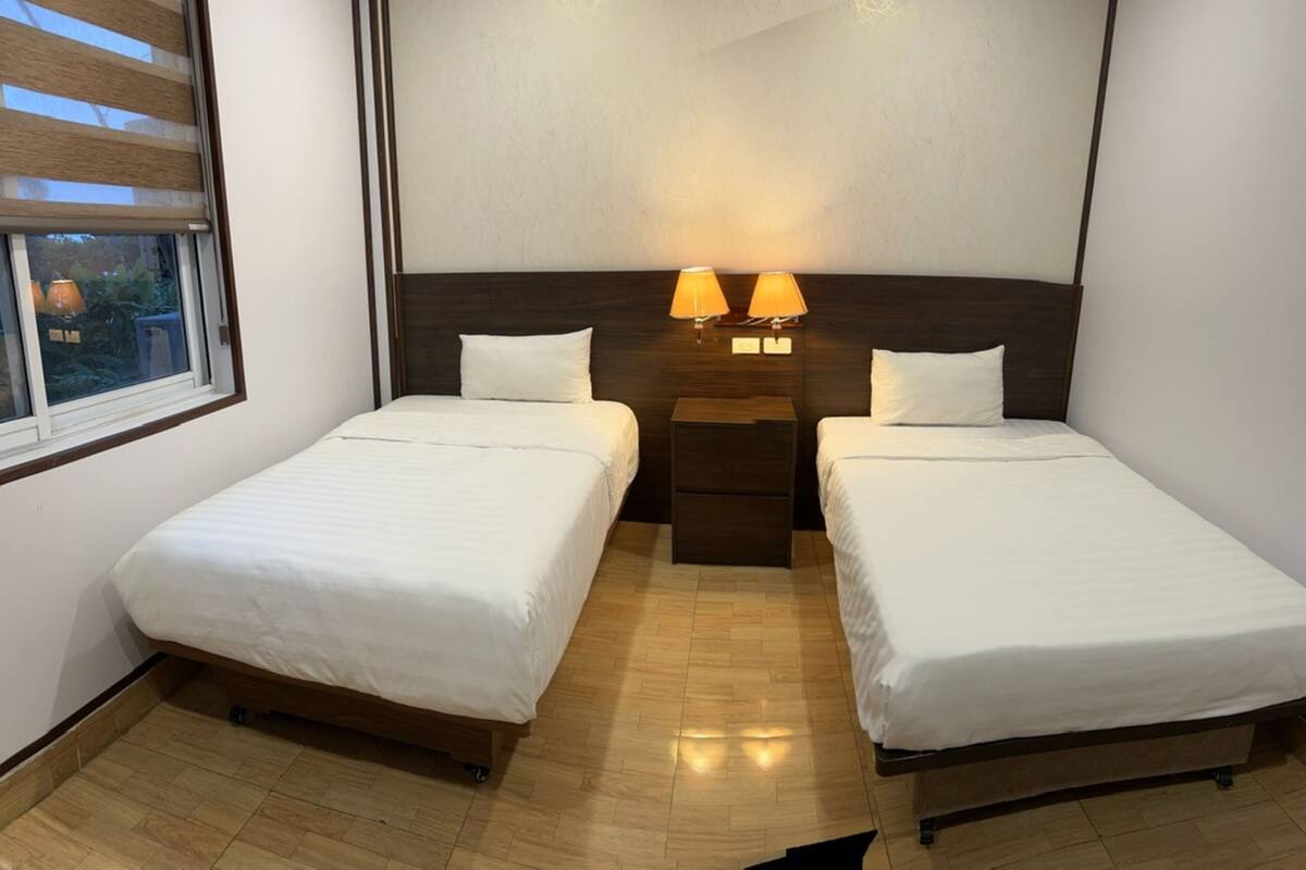 A hotel room with two single beds, a nightstand with lamps, and a window with blinds.