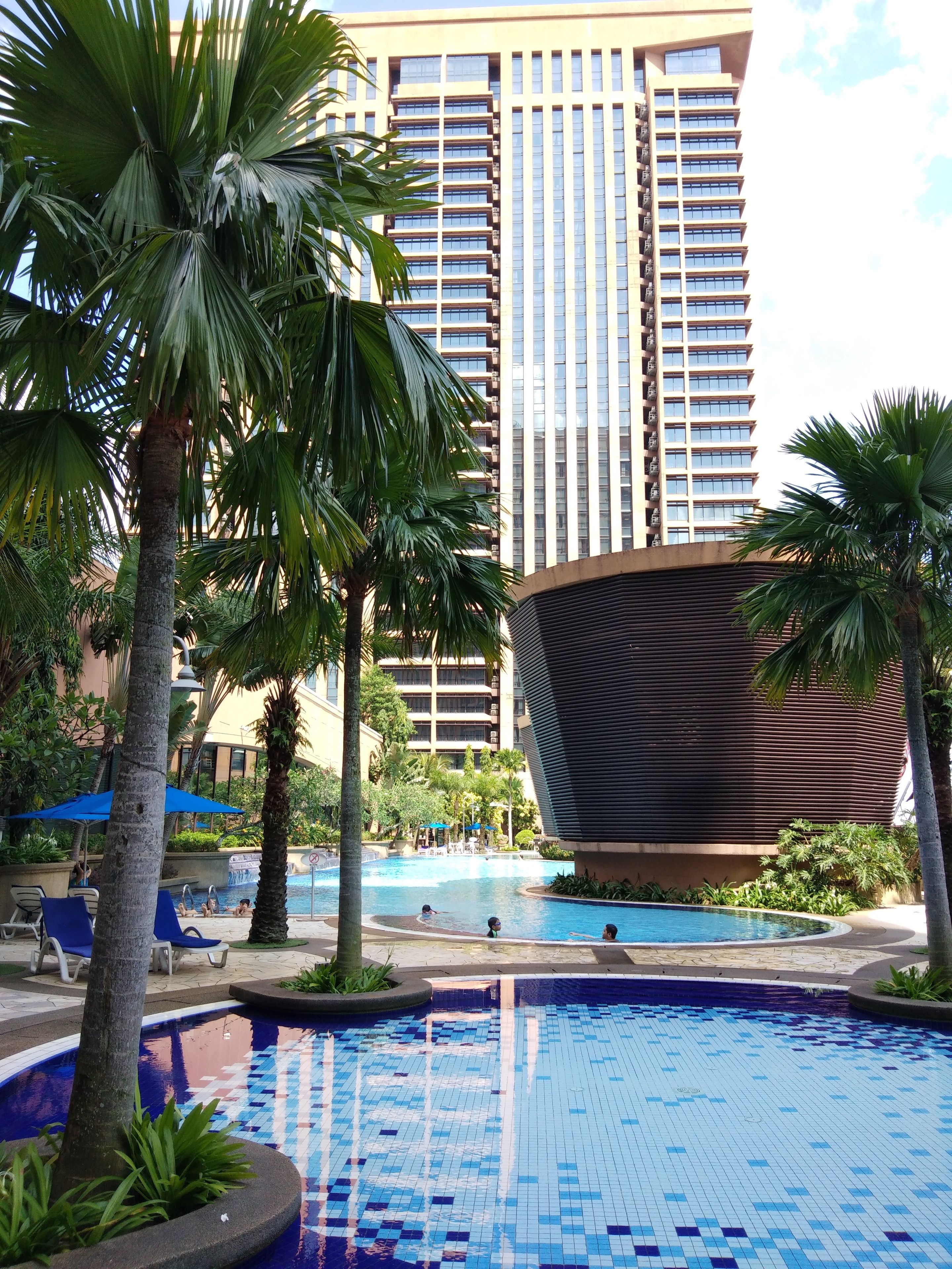 A hotel pool area with a large building in the background, palm trees, and lounge chairs.