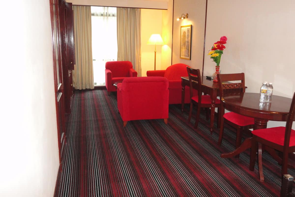 A hotel room with a dining area featuring a wooden table, red chairs, and a red sofa. There is a lamp, a vase with flowers, and a painting on the wall.
