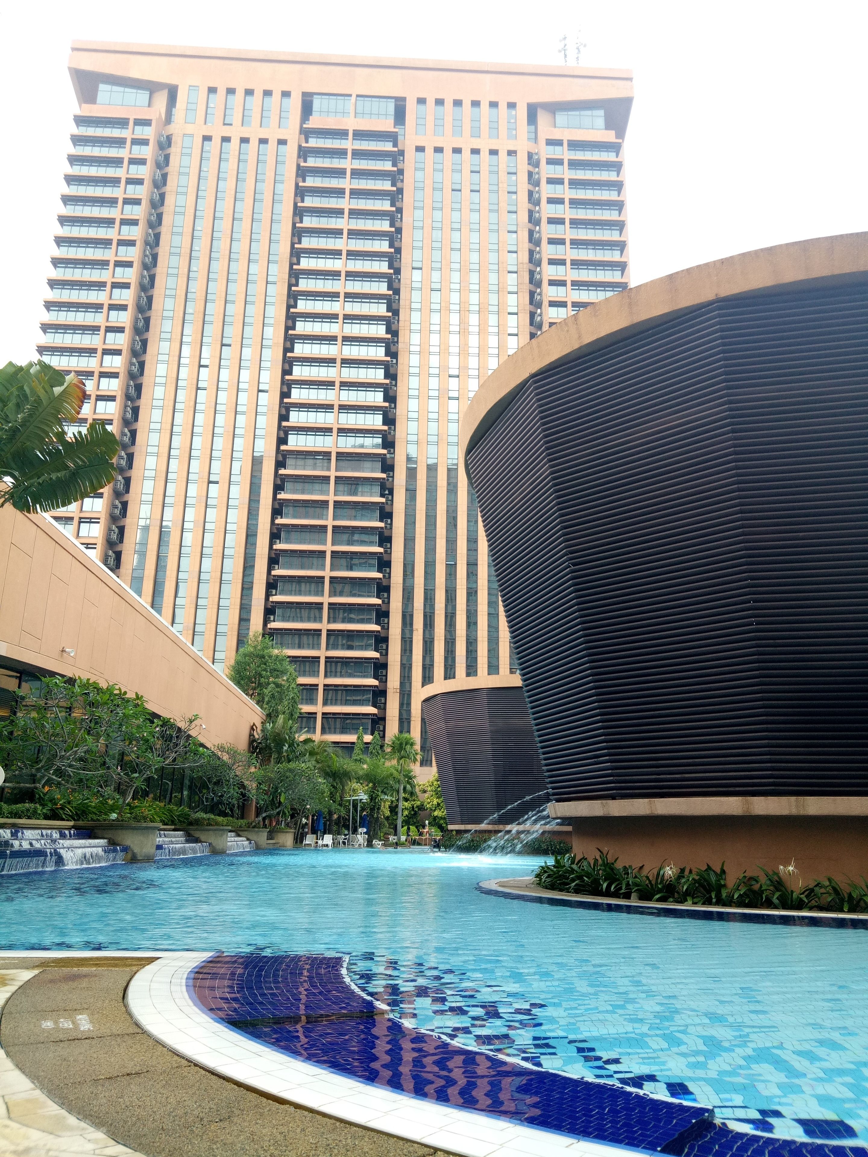 A swimming pool with a tiled edge, surrounded by greenery and a modern building with a curved facade in the background.