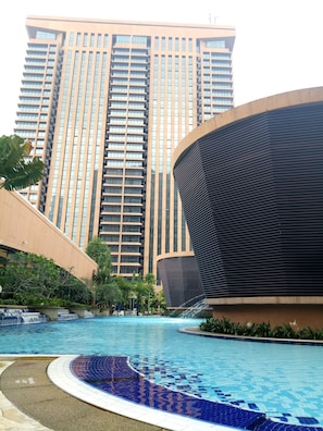 A swimming pool with a tiled edge, surrounded by greenery and a modern building with a curved facade in the background. - KL Times Square Residence (Kuala Lumpur)