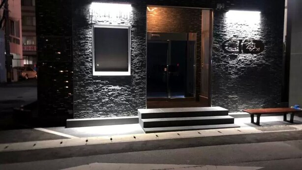 A building with a dark facade, a glass door, and steps leading to the entrance. - Capsule Hotel 2nd BOX (Naha)