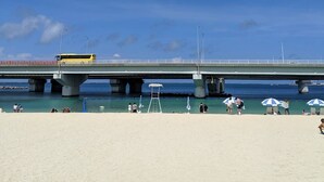 A beach with a bridge overhead, clear blue sky, and people relaxing. - Capsule Hotel 2nd BOX (Naha)