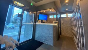 A hotel reception area with a modern design, including a reception desk, a flat-screen TV, and a large window offering a view of the street. - Capsule Hotel 2nd BOX (Naha)