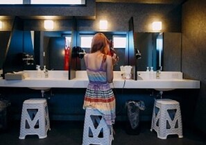 A woman standing at a counter with a mirror above it, in a room with a dark floor and walls, and two stools in front of the counter.