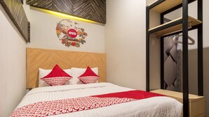 Un lit bien fait, avec une tête de lit en bois, une couvre-lit à motifs rouges et blancs, et des coussins assortis. On y trouve une petite table de chevet en bois, dotée d’un tiroir, et un miroir placé au-dessus.