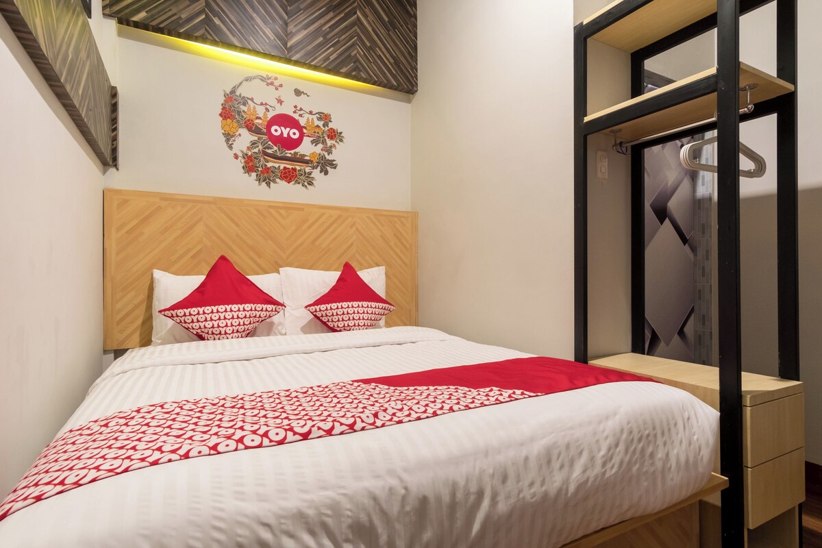 A neatly made bed with a wooden headboard, a red and white patterned bedspread, and matching pillows. There is a small wooden nightstand with a drawer and a mirror above it.