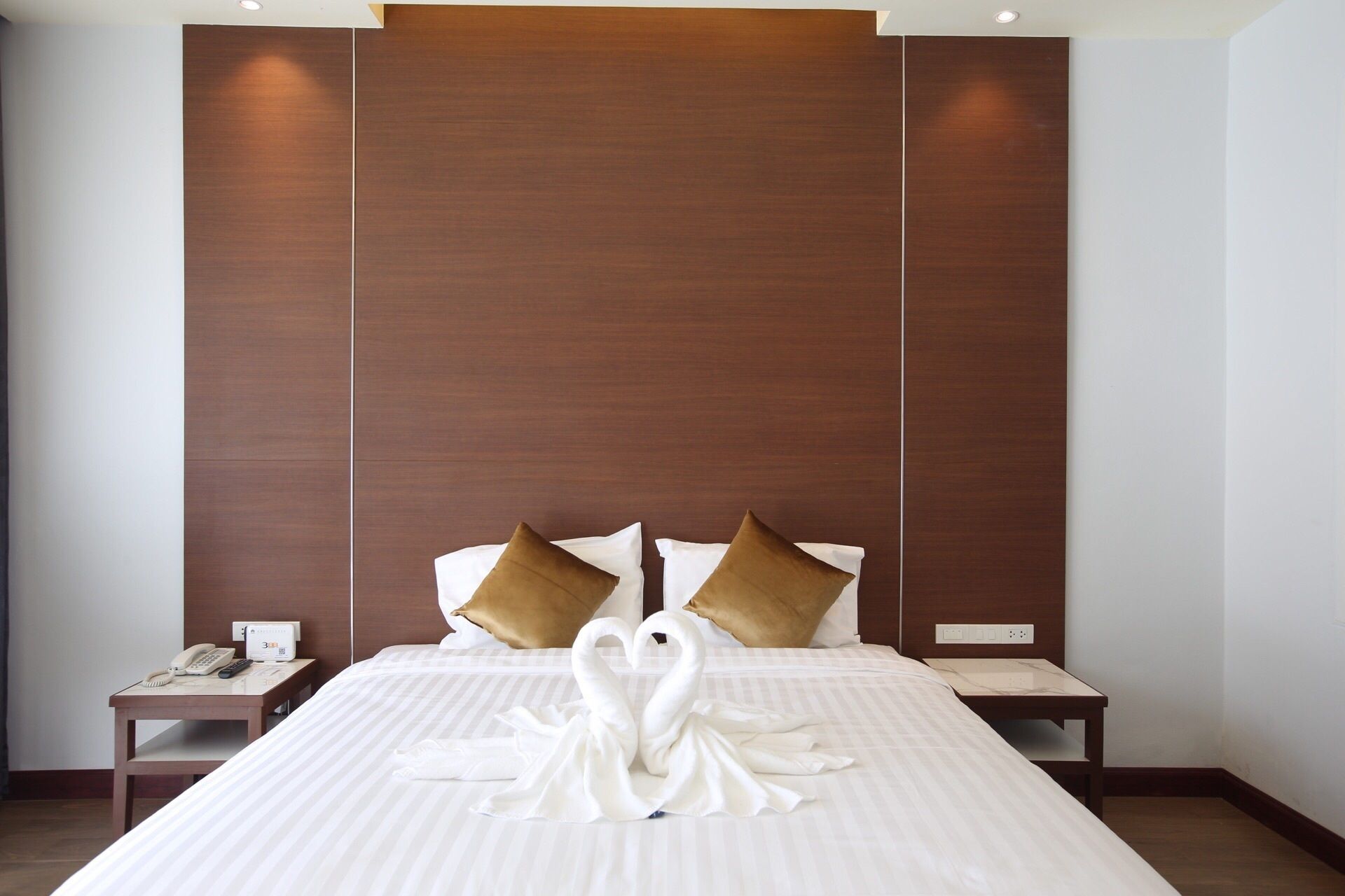 Habitación de hotel con una cama grande, dos almohadas, una mesita de noche con teléfono y una decoración de toallas en forma de cisne sobre la cama.