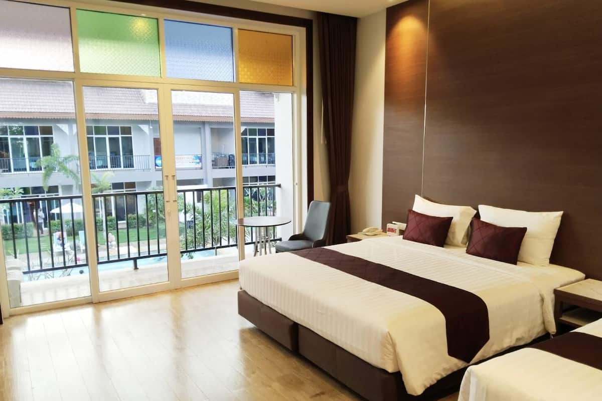Een hotelkamer met een groot bed, een tafeltje en een stoel. Er is een balkon met uitzicht op een gebouw en een zwembad.
