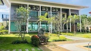 Un bâtiment moderne doté de plusieurs unités de climatisation, d’une galerie couverte et d’un jardin bien entretenu avec diverses plantes et un petit espace piscine.
