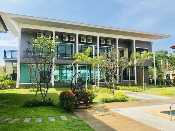 Un bâtiment moderne doté de plusieurs unités de climatisation, d’une galerie couverte et d’un jardin bien entretenu avec diverses plantes et un petit espace piscine.