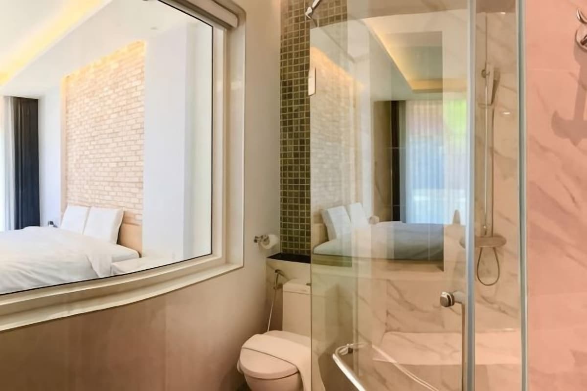Un baño moderno con cabina de ducha de vidrio, inodoro y un espejo grande que refleja una cama y una pared de ladrillo.