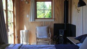 Une pièce en bois avec une grande fenêtre, une chaise, un lit et un poêle à bois.