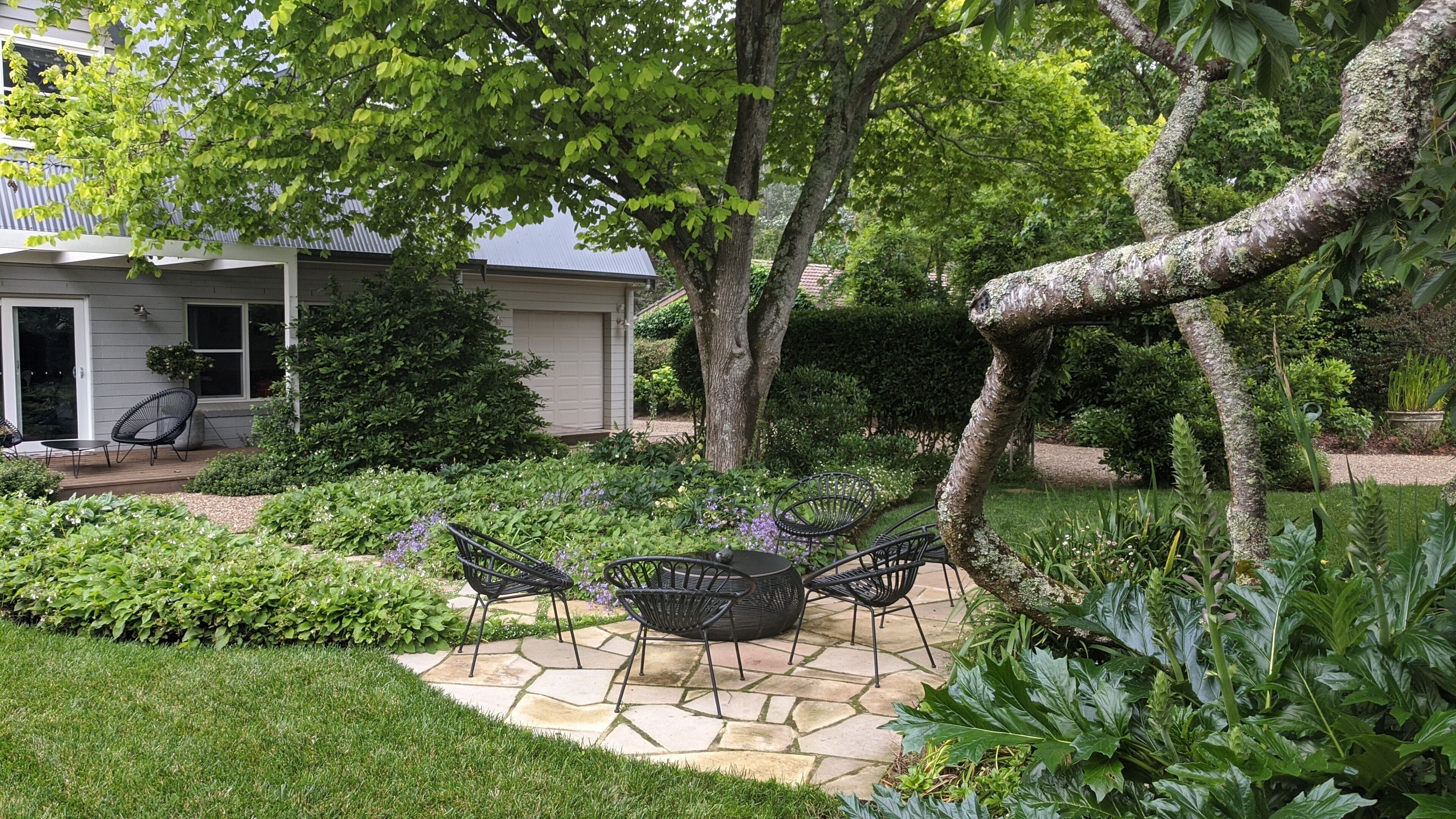 Une terrasse de jardin avec un sol en pierre, entourée d’une végétation luxuriante et de différentes espèces de plantes.