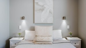 Une chambre à coucher comprenant un lit, deux tables de chevet, une commode, une lampe fixée au mur et un tableau encadré.