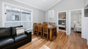 Un salon moderne avec un canapé en cuir noir, une table à manger en bois et des chaises. On y trouve une grande fenêtre avec des stores, un plancher en bois et, au fond, un lit superposé.